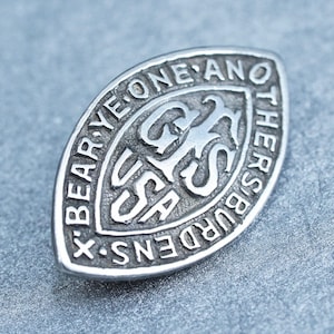 Puede incluir: Un pin ovalado de color plateado con el texto "X BEAR YE ONE ANO THERS BURDENS X" alrededor del borde. El centro presenta las letras "GS" y "USA". El pin es de metal y tiene una superficie ligeramente texturizada.