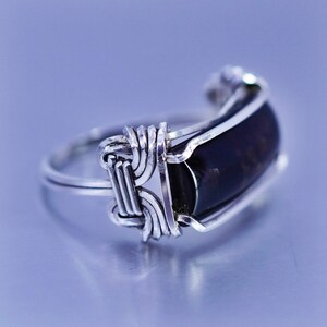 May include: A silver ring with a dark, rectangular stone. The stone is set in a silver frame and secured with intricate wire wrapping. The ring has a polished finish and a simple band.
