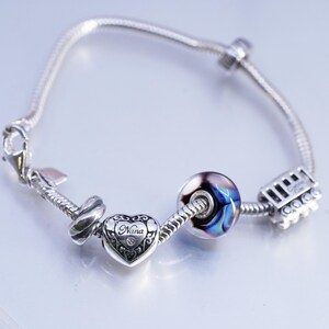 Puede incluir: Una pulsera de plata con varios abalorios. Los abalorios incluyen un corazón con la palabra "Nana", una cuenta de vidrio con remolinos azules y morados, y un pequeño tren. La pulsera está sobre un fondo claro.