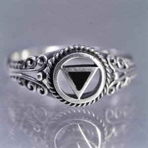 May include: A silver ring featuring a central design of a triangle within a circle. The ring has a rope-like border around the central design and ornate scrollwork on the band. The triangle is filled with a dark material, creating a contrast with the silver.