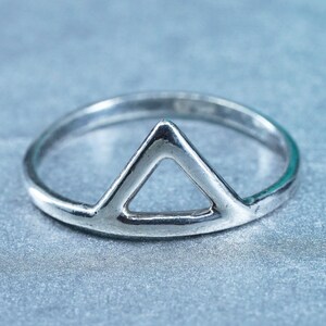 May include: A silver ring with a triangular design. The ring features a simple, open triangle shape at the center, set on a thin band. The ring is made of a shiny, metallic material and is displayed against a light blue background.