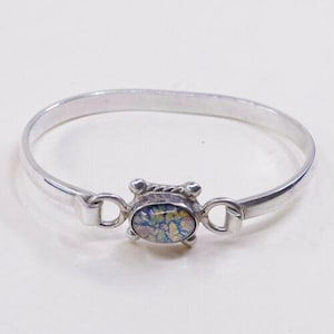May include: A silver-toned bangle bracelet with an oval-shaped, dichroic glass centerpiece. The glass displays a mix of blue, yellow, and green colors. The bracelet has a simple, elegant design with a clasp closure.