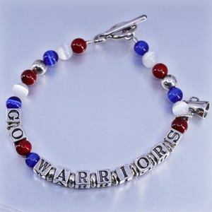 May include: Silver-toned bracelet with red, white, and blue beads. The bracelet spells out "GO WARRIORS" with silver lettered cubes. A silver charm shaped like a megaphone is attached to the bracelet.