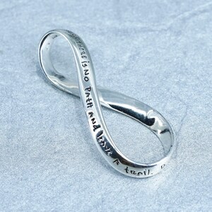May include: A silver-colored infinity symbol pendant with the inscription "there is no path and I have a trail." The pendant is made of a shiny, reflective metal and is displayed on a gray surface.