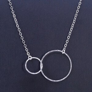 May include: A silver necklace with two interlocking hammered rings. The larger ring is approximately 3.8 cm in diameter, and the smaller ring is about 1.9 cm. The necklace chain is a delicate link style, set against a dark background.