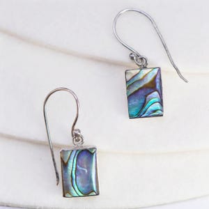 May include: A pair of rectangular drop earrings with iridescent abalone shell inlays. The earrings have silver-colored hooks and frames, showcasing the natural blue, green, and purple hues of the shell. The earrings are displayed on a white surface.