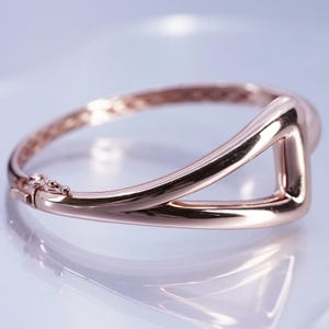 May include: Rose gold bangle bracelet with a geometric design. The bracelet has a hinged clasp and a triangular cutout. The polished metal reflects light, creating a shiny appearance. The jewellery is displayed on a reflective surface.