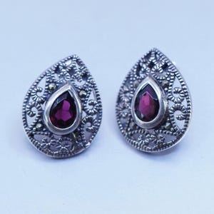 May include: Pair of silver-toned teardrop earrings. Each earring features a deep red, teardrop-shaped gemstone set in a filigree design. The filigree is accented with small, round, silver-colored details.