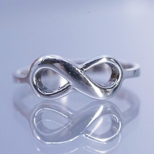 May include: A silver-colored ring featuring an infinity symbol design. The ring is highly reflective, with a smooth, polished surface. The infinity symbol is the focal point, with a continuous loop shape. The ring is likely made of a metal such as sterling silver.