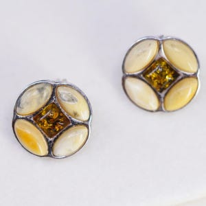 May include: A pair of round stud earrings with a silver-tone metal setting. Each earring features four oval-shaped, cream-colored stones surrounding a central, diamond-shaped section of amber-colored stones. The earrings are set against a white background.
