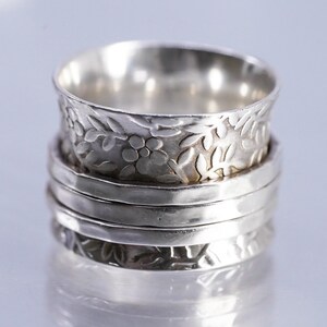 May include: A silver spinner ring featuring a wide band with an embossed floral and leaf pattern, layered with two smooth, polished silver bands in the center. The metal has a reflective finish with darkened recessed areas to highlight the design.