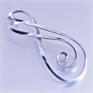 May include: A polished silver-toned pendant in an infinity symbol design. The pendant features a flowing, looped shape with a spiral detail at the bottom. The metal reflects light, creating highlights and shadows.