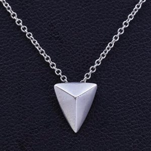 May include: A silver-colored necklace with a geometric triangular pendant. The pendant is a solid, three-dimensional pyramid shape. The chain is a delicate, linked design. The background is a dark, textured surface.