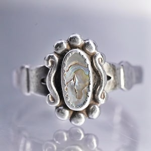 May include: A silver ring with an oval abalone shell center stone. The ring has a decorative border of small silver beads and scrollwork. The band features rectangular accents on each side of the ring.