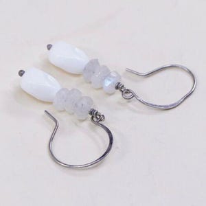 May include: A pair of dangle earrings featuring white, oval-shaped beads and small, irregular, light-colored stones. The earrings have silver-colored, curved ear wires. The design is simple and elegant, suitable for everyday wear or special occasions.