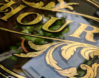Pode incluir: Uma placa de folha de ouro com a palavra "HOUR" em letras pretas. A placa tem uma borda decorativa e um fundo de folha de ouro.