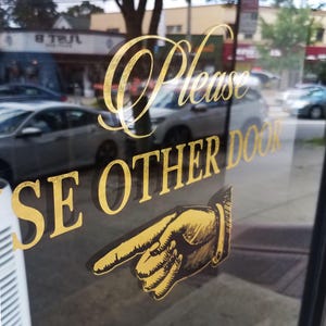 May include: A glass door with gold lettering that reads "Please Use Other Door." A gold illustration of a hand pointing to the right is below the text. Cars and buildings are visible through the glass.