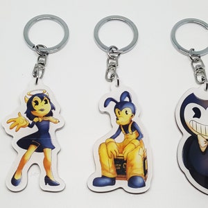 May include: Four keychains featuring cartoon characters. Each keychain has a silver metal ring and a chain. The characters include a smiling devil, a female angel, a dog-like character, and a smiling character with a bow tie.