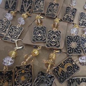 Puede incluir: Pendientes dorados con diseños de cartas del tarot negras y detalles dorados, con cuentas colgantes. Los pendientes muestran varias ilustraciones de cartas del tarot, incluyendo la carta "La Fuerza" con un león y otros símbolos.