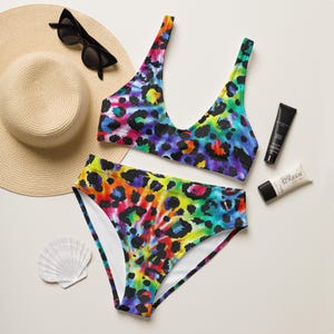 May include: A two-piece bikini set featuring a vibrant rainbow tie-dye pattern with black leopard spots. The set includes a scoop-neck top and high-waisted bottoms, accompanied by a straw hat, black sunglasses, two tubes of sunscreen, and a seashell.