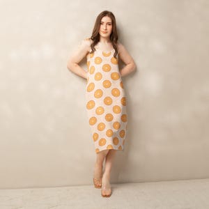 May include: A knee-length dress with a light beige background and a repeating pattern of orange citrus fruit slices. The dress has thin straps and a V-neckline. The model is wearing clear sandals.