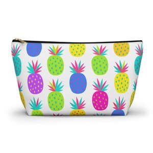 Può includere: Una pochette bianca con cerniera caratterizzata da un motivo ripetuto di ananas stilizzati in colori vivaci, tra cui giallo, verde lime, blu brillante e rosa acceso. Ogni ananas ha un ciuffo di foglie multicolore nei toni del verde acqua, rosa e verde.