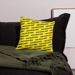 May include: A yellow throw pillow with the text "YOUR DESIGN HERE!" in black repeated across the surface. The pillow is placed on a dark gray sofa with a green blanket.