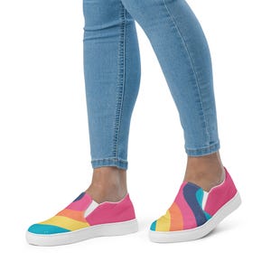 May include: Slip-on sneakers with a vibrant rainbow design. The shoes feature a white sole and a bright pink upper. The rainbow pattern includes shades of blue, yellow, orange, and pink, creating a colorful and eye-catching look.