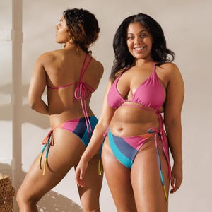 May include: Two women wearing matching pink and multicolored bikinis. The bikini tops are a solid pink, while the bottoms feature a striped design in shades of pink, blue, and yellow. The swimwear is tied with pink strings.