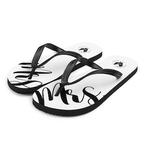May include: White flip-flops with black straps and soles. The word "Mrs" is written in a large, elegant black script across the top of the flip-flops. A small black graphic is on the top of each flip-flop.