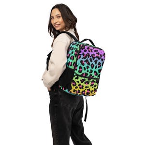 Puede incluir: Una mochila colorida con un estampado de leopardo vibrante. La mochila presenta un degradado de colores, incluyendo rosa, morado, verde y amarillo. Tiene bolsillos laterales y correas negras. Ideal para llevar libros y útiles escolares.