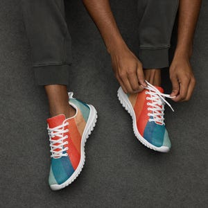 May include: Colorful sneakers with a retro striped design in red, orange, tan, and blue. The shoes have white laces and soles. The person is tying the laces of the sneakers.