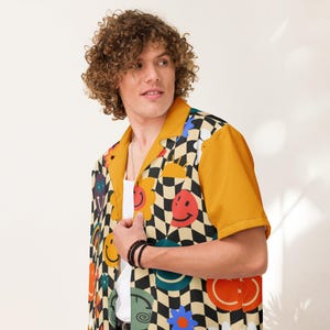 Puede incluir: Una camisa de manga corta con un estampado de tablero de ajedrez y caras sonrientes. La camisa tiene mangas y cuello de color amarillo mostaza. El diseño de la camisa incluye flores y otras formas coloridas. La camisa se lleva sobre una camiseta interior blanca.
