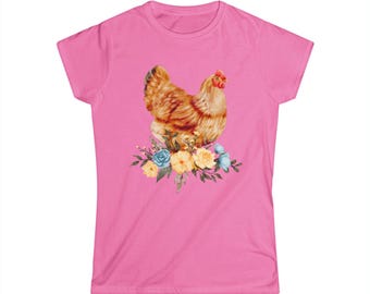 May include: A pink t-shirt featuring a watercolor illustration of a hen perched atop a floral arrangement. The hen is brown and tan with a red comb. The flowers are blue and yellow.