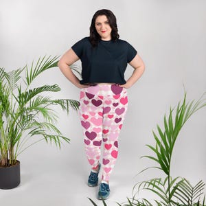 May include: Plus-size leggings featuring a pattern of pink and purple hearts on a white and pink checkered background. The leggings are paired with a dark navy blue t-shirt. The model is wearing blue sneakers.