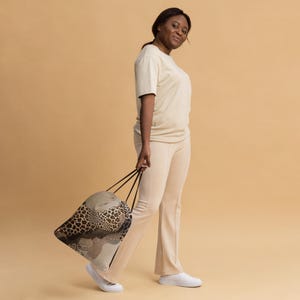 May include: A beige drawstring bag with a leopard print design is held by a person. The bag features a mix of brown, tan, and cream colors. The person is wearing a beige t-shirt and pants.
