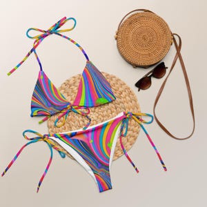 May include: A colorful bikini set with a vibrant, swirling pattern in shades of blue, pink, yellow, and green. The set includes a halter top and tie-side bottoms. A round woven bag, sunglasses, and a straw mat are also in the image.