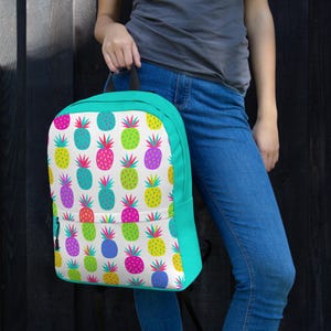 May include: A backpack featuring a white background with a repeating pattern of colorful pineapples in shades of pink, yellow, green, purple, and blue. The backpack has solid teal side panels, a top handle, and a front zippered pocket.