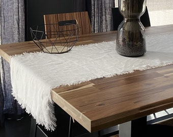May include: A white linen table runner with frayed edges sits on a wooden table with a black metal wire bowl and a glass vase filled with dried lavender.