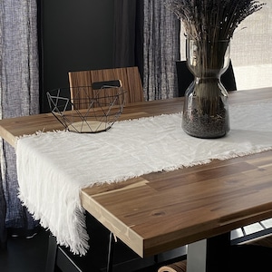 May include: A white linen table runner with frayed edges sits on a wooden table with a black metal wire bowl and a glass vase filled with dried lavender.