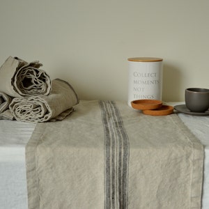 Könnte beinhalten: Eine weiße Keramikdose mit einem Holzdeckel und dem Text "Collect Moments, Not Things". Die Dose steht auf einem Tisch mit einem Tischläufer aus Leinen mit schwarzen Streifen. Neben der Dose befinden sich zwei braune Untersetzer. Eine braune Keramik-Kaffeetasse und eine Untertasse stehen auf dem Tisch.