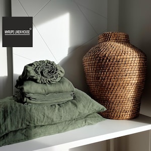 May include: A stack of green linen bedding, including a fitted sheet, flat sheet, and pillowcases, on a white shelf with a woven basket vase. The text "MYRUPE LINEN HOUSE" is on a black label.