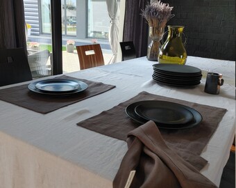 May include: A white tablecloth with two brown placemats and two black plates. A brown linen napkin is folded on the table. The table is set for a formal dinner.