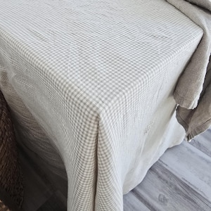 Puede incluir: Un mantel a cuadros beige y blanco cubre una mesa. La tela cae sobre los bordes, con un paño doblado encima. El patrón es un diseño de cuadrícula pequeño y repetitivo, que crea una estética clásica y neutra.