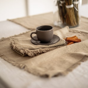 May include: A brown ceramic coffee cup and saucer on a set of two beige linen placemats with frayed edges. The placemats are on a white tablecloth.