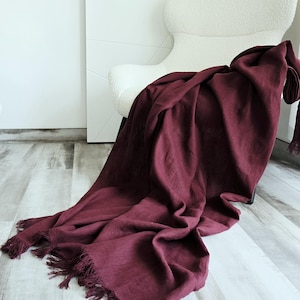 Puede incluir: Una manta burdeos drapeada con bordes con flecos descansa sobre un sillón blanco texturizado. La tela de la manta es suave y parece ser lino o un material similar. El fondo incluye una pared blanca y un suelo de madera clara.