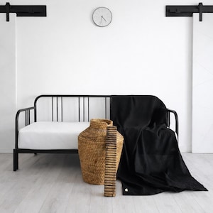 Peut inclure: Un canapé-lit en métal noir avec un matelas blanc et une couverture noire. Un grand panier tissé et une pièce décorative en bois se trouvent à côté du lit. La pièce a des murs blancs, un sol en bois clair et une horloge simple.