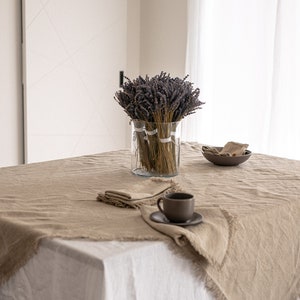 May include: A light brown linen tablecloth with frayed edges, a white tablecloth underneath, a vase of dried lavender, a cup and saucer, and a small bowl on a white table.
