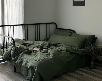 Könnte beinhalten: Ein grüner Leinen-Bettbezug und Kissenbezüge auf einem schwarzen Metallbettgestell. Das Bett steht in einem Raum mit einer weißen Wand und einem weißen Leinenvorhang. Der Text "MYRUPE LINEN HOUSE" ist an der Wand sichtbar.