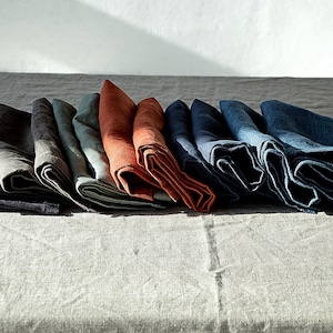 May include: A set of folded linen napkins in various shades of blue, green, and orange. The napkins are arranged in a row on a light grey linen tablecloth.